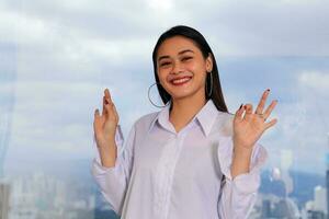 Young beautiful modern Malay woman relax wave hand to camera next to window cityscape cloudy blue sky photo