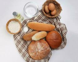 Freshly baked bread loaf bun roll round long mix verity wrapped in checkered kitchen fabric napkin towel wheat flower oil water salt eggs over white background photo