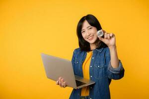 Happy young asian woman wearing yellow t-shirt denim shirt holding digital crypto currency coin and laptop isolated on yellow background. Digital currency financial concept. photo