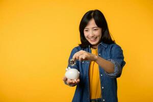 Happy young asian woman wearing yellow t-shirt denim shirt saving digital crypto currency to piggy bank isolated on yellow background. Digital money saving investment concept. photo