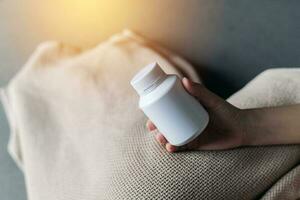 pain ill sick young female adult person hand hold pill medicine bottle over a blanket on bed. disease, health, medical, treatment pharmacy, vitamin, supplement take care concept. photo