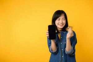 Happy young asian woman wearing yellow t-shirt denim shirt holding digital crypto currency coin and smartphone isolated on yellow background. Digital currency financial concept. photo