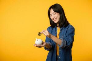 Happy young asian woman wearing yellow t-shirt denim shirt saving digital crypto currency to piggy bank isolated on yellow background. Digital money saving investment concept. photo