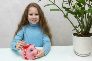 pequeño niña con largo pelo pone dinero en un cerdito banco foto