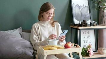 mulher sentado às cama às lar, tendo café da manhã e usando Smartphone. jovem milenar mulher fazendo dela manhã rotina às a quarto video