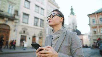 Woman with glasses wearing a coat walking down an old street and using smartphone video