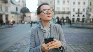 Woman with glasses wearing a coat walking down an old street and using smartphone video