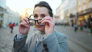 portrait de une caucasien femme dans une manteau permanent dans le milieu de le vieux ville carré. elle regards à le caméra et met sur des lunettes video