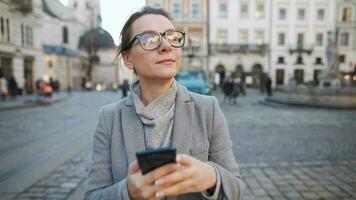 Frau mit Brille tragen ein Mantel Gehen Nieder ein alt Straße und mit Smartphone video