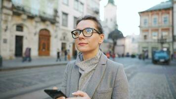 vrouw met bril vervelend een jas wandelen naar beneden een oud straat en gebruik makend van smartphone video