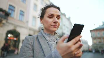 Frau mit Brille tragen ein Mantel Gehen Nieder ein alt Straße und mit Smartphone video