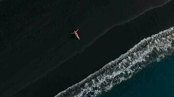 Aerial view of a woman in a red swimsuit lying on a black beach on the surf line. Coast of the island of Tenerife, Canary Islands, Spain video