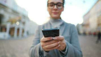 mujer con lentes vistiendo un Saco caminando abajo un antiguo calle y utilizando teléfono inteligente video