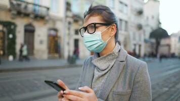 mujer en protector médico máscara caminando abajo un antiguo calle y utilizando teléfono inteligente video