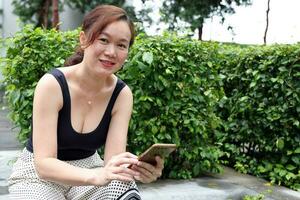 beautiful attractive middle age southeast asian woman sitting relaxing reading texting smart phone in urban green garden background photo
