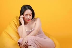 Beautiful young south east Asian woman sit on a yellow orange beanbag seat color background relax rest think emotion imagine expression pose sad photo