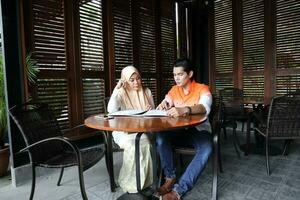 Young asian malay man woman with headscarf sit at rustic cafe table talk discuss business study read photo