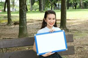 Young asian chinese woman outdoor on park bench hold whiteboard copy text space smile look point photo