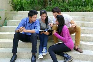 group of Asian young man woman friend student colleagues sting on stairs talk mingle discuss study book note photo