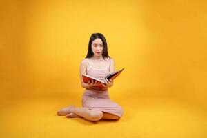 Beautiful young south east Asian woman sit down on floor yellow orange color background read study book magazine pose fashion style elegant beauty mood photo