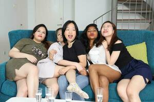 joven asiático mujer grupo hablar chisme charla canta bebida salud fiesta divertido disfrutar gracioso caras expresión en azul vivo habitación sofá foto