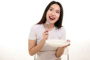 hermosa joven sur este asiático mujer fingir interino posando participación vacío tenedor cuchara blanco plato en mano comer gusto Mira ver blanco antecedentes sonrisa contento comida en boca imagina foto