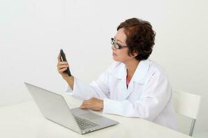 Elderly asian Chinese female doctor on white background wearing white apron reading text on phone using laptop computer photo