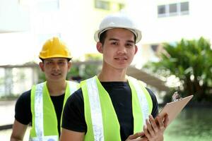 South East Asian young Malay Chinese man woman safety helmet vest formalwear stand at construction site blueprint plan clipboard look at camera photo