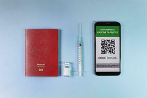 Digital international vaccine passport smart phone screen qr code app coronavirus covid 19 immunization red passport book small bottle disposable medical syringe needle on light sky blue background photo