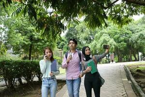 joven asiático malayo chino hombre mujer al aire libre parque caminar estar ordenador portátil archivo libro mochila utilizar mano teléfono selfie foto