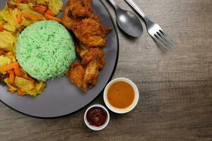 Steamed green rice spiced deep fried chicken cabbage carrot vegetable warung nasi ayam goreng on dark grey plate wooden background gravy sambal sauce fork spoon photo