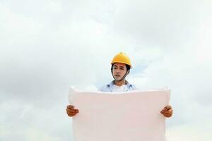 asiático hombre malayo trabajador ingeniero administración difícil la seguridad sombrero casco a construcción sitio leyendo viendo plan azul cielo foto