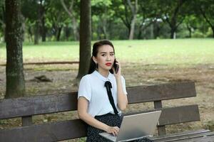 Young asian chinese woman outdoor on park bench use laptop computer think read look happy wonder smile front talk on phone photo