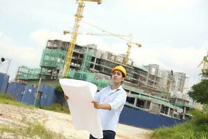 asiático hombre malayo trabajador ingeniero administración difícil sombrero la seguridad casco a construcción sitio leyendo viendo plan Plano foto