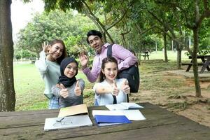 Young asian malay chinese man woman outdoor on park bench table study talk discuss point laptop file book happy pose look forward thumbs up ok hand sing wave photo