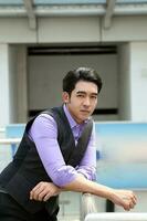 Southeast Asian young office business man wearing vest lean on railing stand look at camera confident expression outdoor premises photo