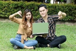 Young Asian Malay man woman outdoor green park sit on grass use study discuss laptop thumbs down hand sign photo