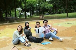 Young asian malay chinese man woman outdoor park sit on ground study talk discuss point laptop file book backpack mingle fun thumbs up photo