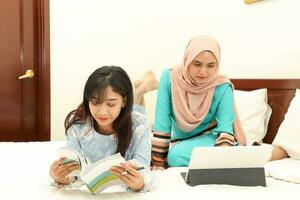 Two young Asian Malay Muslim woman wearing headscarf baju kurung dress at home on bed study work on computer tab book notebook pen read write relax photo