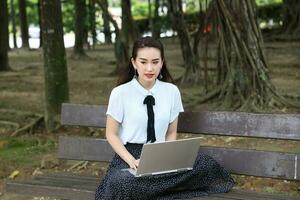 Young asian chinese woman outdoor on park bench use laptop computer think read look happy wonder smile look forward photo