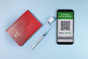 Digital international vaccine passport smart phone screen qr code app coronavirus covid 19 immunization red passport book small bottle disposable medical syringe needle on light sky blue background photo