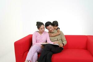 South East Asian young couple father mother daughter parent girl child on red sofa read write study book on white background photo