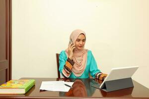 young Asian Malay Muslim woman wearing headscarf at home office student sitting at table talk mingle look at computer pone book document study discuss talk smile happy photo