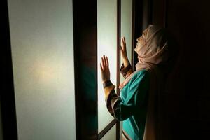 joven asiático malayo musulmán mujer vistiendo Pañuelo baju Kurung vestir a hogar en frente ventana estado animico ligero en cara oscuro antecedentes pacífico contenido calma tranquilo descanso canal preguntarse foto