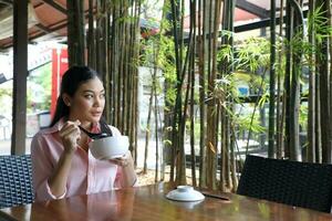 Young asian malay woman sit at rustic cafe table eat with chinese spoon from soup noodle bowl look outside photo