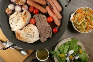 Mix meet chicken beef steak sausage tomato onion garlic fry grill oil metal fry pan colourful pasta green salad with cheese olive mint basil pesto sauce board knife fork spoon rustic wooden table photo