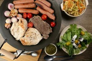 Mix meet chicken beef steak sausage tomato onion garlic fry grill oil metal fry pan colourful pasta green salad with cheese olive mint basil pesto sauce board knife fork spoon rustic wooden table photo