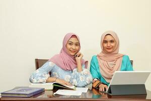 Two young Asian Malay Muslim woman wearing headscarf at home office student sitting at table talk mingle look at computer book document study discuss smile happy photo