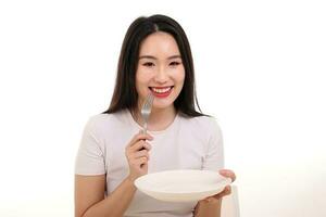 hermosa joven sur este asiático mujer fingir interino posando participación vacío tenedor cuchara blanco plato en mano comer gusto Mira ver blanco antecedentes sonrisa contento foto