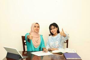 Two young Asian Malay Muslim woman wearing headscarf at home office student sitting at table talk mingle computer book document study thumbs up sign look at camera photo